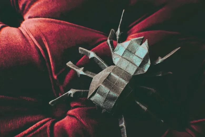 a large , complex origami spider crawling on a red velvet cushion . the spider is black with a teal sparkle